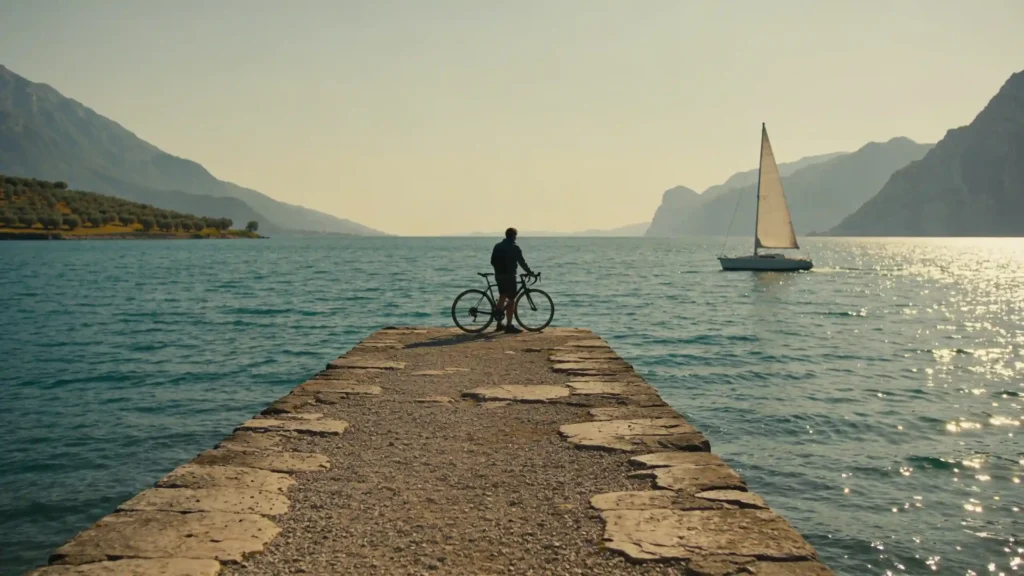 Un cycliste observe un voilier sur un vaste lac italien bordé de montagnes.